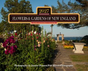 Signboard advertising a 2027 'Flowers & Gardens of New England' calendar, set among colorful garden flowers by a waterfront path.
