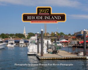 Boat marina with several yachts docked, under a large Rhode Island Scenic Engagement Calendar sign overhead.
